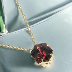 May include: A gold necklace with a large, faceted, reddish-brown gemstone pendant. The pendant is hexagonal and set in a gold bezel. The chain is delicate and gold, resting on a light blue surface.
