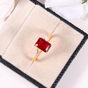 May include: A gold ring featuring a large, rectangular, deep red gemstone as the centerpiece. Two smaller, pear-shaped white gemstones flank the main stone. The band is gold with small, sparkling accents. The ring is presented in a black and beige jewelry box.