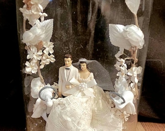 Op de afbeelding: Een vintage bruidstaarttopper in een glazen stolp. De topper toont bruid en bruidegom beeldjes, omringd door witte bloemdecoraties. De basis van de stolp is versierd met heldere kralen, wat een elegante presentatie creëert.