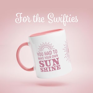 Könnte beinhalten: Weißer Keramikbecher mit rosa Innenraum und Henkel. Der Becher zeigt den Text "YOU HAD TO MAKE YOUR OWN SUN SHINE" in Rosa, mit einem Sonnenstrahl-Design. Die Worte "For the Swifties" stehen oben.