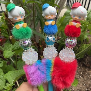 May include: Three decorative pens featuring cartoon duck figures with different colored hats. Each pen has a fluffy pom-pom base in green, blue, and red, with iridescent and floral bead accents. The pen barrels are painted in coordinating colors.