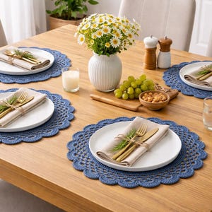 May include: A wooden dining table set for a meal. Each place setting includes a white plate, linen napkin, and gold silverware on a blue lace placemat. A vase of daisies, grapes, and snacks are also on the table.