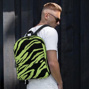 Puede incluir: Una mochila con un estampado de cebra en negro y verde neón. Tiene un bolsillo frontal con cremallera y correas negras. El fondo es un panel de madera oscura.