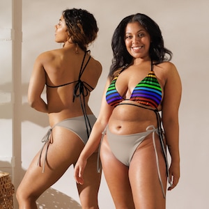 May include: Two women wearing bikinis. One bikini top is a rainbow striped pattern, and the other is black. The bikini bottoms are a solid light gray color. The bikinis have tie closures.