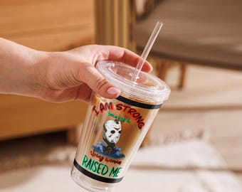 May include: Clear plastic tumbler with a lid and straw. The tumbler features a graphic of a hockey mask and the text "I AM STRONG because a strong woman RAISED ME". The tumbler is filled with a brown liquid.