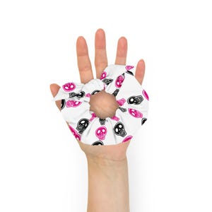 Puede incluir: Coletero blanco con un patrón repetido de calaveras negras y rosas. El accesorio para el cabello se sostiene en una mano sobre un fondo blanco. Las calaveras tienen un diseño de dibujos animados.