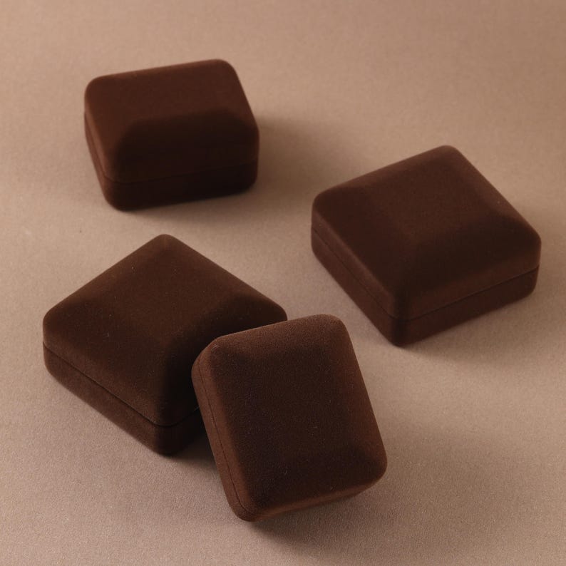 May include: Four square jewelry boxes with a dark brown velvet-like finish. Each box features a beveled lid and a compact, closed design. The boxes are arranged in a cluster, showcasing their uniform shape and deep brown color throughout the set.