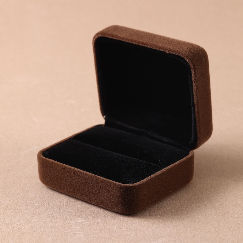 May include: A square jewelry box with a brown velvet exterior and a black velvet interior lining. The box is open, featuring a single slot designed for holding a ring, with a hinged lid that stands upright against the base of the container.