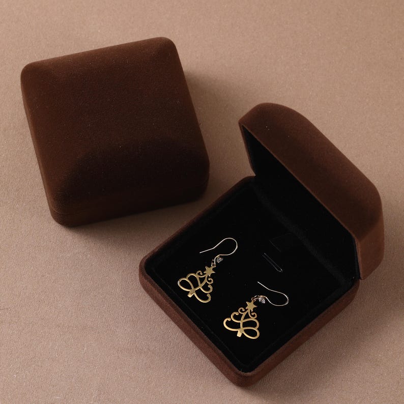 May include: A pair of gold-toned dangle earrings shaped like stylized Christmas trees with a star at the top, presented inside an open, dark brown velvet jewelry box with a matching closed lid resting nearby on a flat surface.