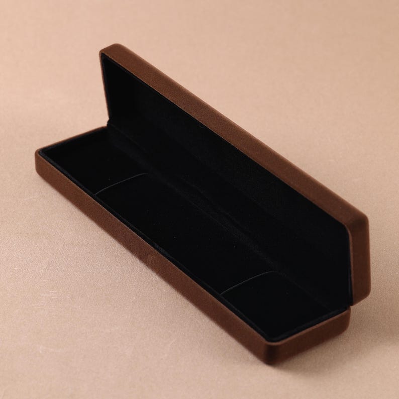 May include: A rectangular jewelry box with a brown exterior and a black interior lining. The box is open, revealing a deep, empty compartment designed for storage. The exterior has a soft, matte finish, while the interior is lined with fabric.