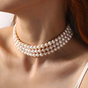 May include: A three-strand pearl choker necklace. The necklace features multiple rows of round, creamy white pearls. The necklace is worn around the neck, showcasing the elegant design and classic style of the jewelry.