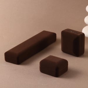 May include: Three dark brown velvet jewelry boxes of varying shapes and sizes. One box is long and rectangular, while the other two are smaller and square-shaped. The boxes have a soft, matte texture and are closed, showing no interior details.