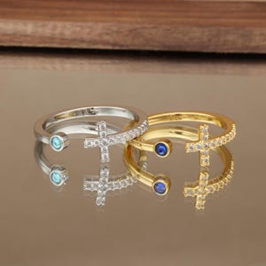 May include: Two open-style rings, one silver-toned and one gold-toned, each featuring a cross-shaped charm encrusted with clear stones on one end and a single round-cut coloured stone on the other, with clear stones lining the band.