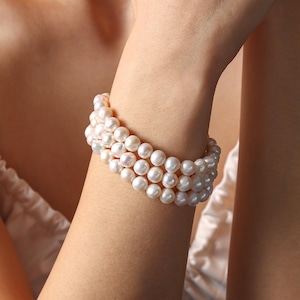 Può includere: Un braccialetto di perle a tre fili. Il braccialetto presenta perle bianche rotonde infilate insieme. Le perle sono di colore bianco crema e sembrano essere di alta qualità. Il braccialetto è indossato su un polso.