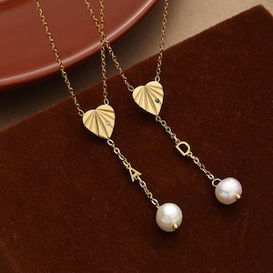 Peut inclure: Deux colliers dorés avec des pendentifs en forme de cœur ornés de stries en éventail. Un pendentif possède une pierre claire, l'autre une pierre verte. Chaque chaîne comporte une lettre, 'A' ou 'D', et se termine par une perle blanche.
