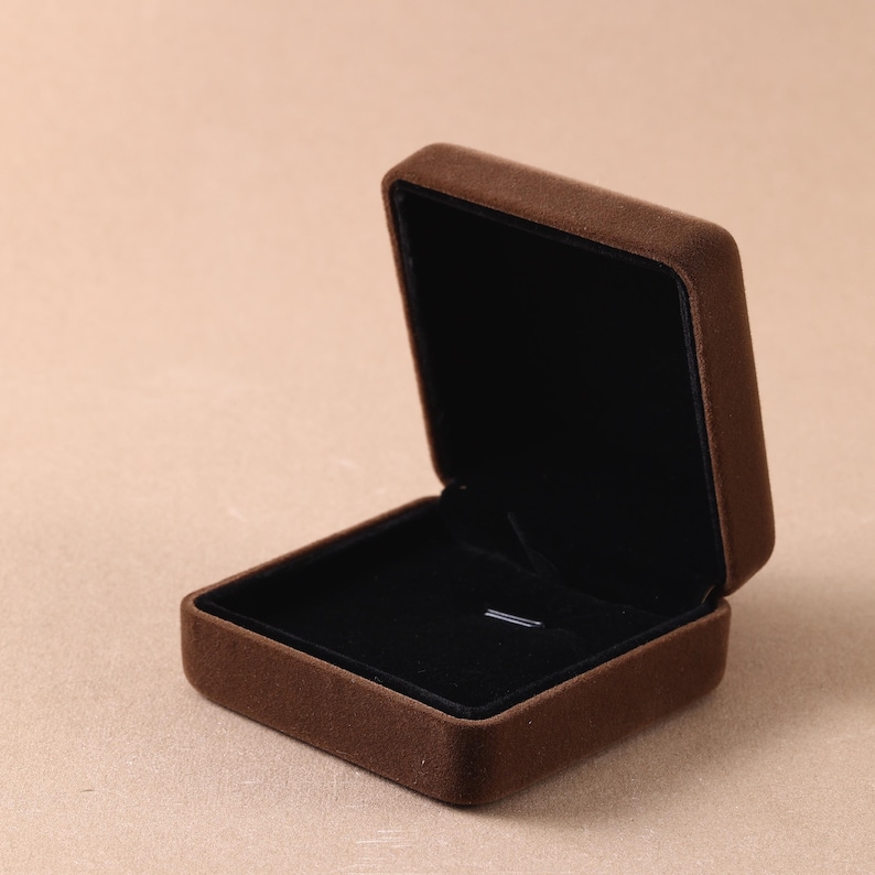 May include: A square jewelry box with a brown velvet exterior and a black velvet interior lining. The box is open, revealing a flat base with a small horizontal slot in the center for securing a piece of jewelry. The lid is hinged at the back.
