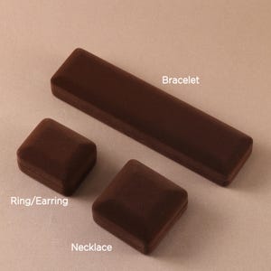 May include: Three dark brown velvet jewelry boxes of varying shapes and sizes. One long rectangular box labeled Bracelet, one square box labeled Ring/Earring, and one square box labeled Necklace. The boxes have a soft, matte, textured finish.