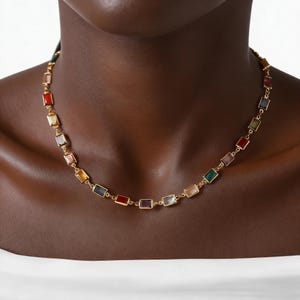 Puede incluir: Un collar dorado con piedras preciosas rectangulares multicolores. Las piedras están engastadas en marcos dorados individuales y unidas para formar una delicada cadena. El collar se lleva alrededor del cuello.