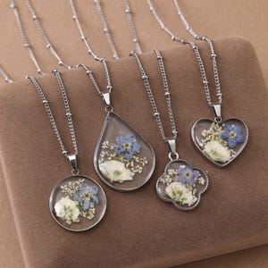 Puede incluir: Cuatro collares plateados con colgantes de resina transparente en formas redonda, de lágrima, de trébol y de corazón. Cada colgante contiene flores blancas y azules prensadas dentro de la resina, suspendidos de delicadas cadenas de metal plateado.