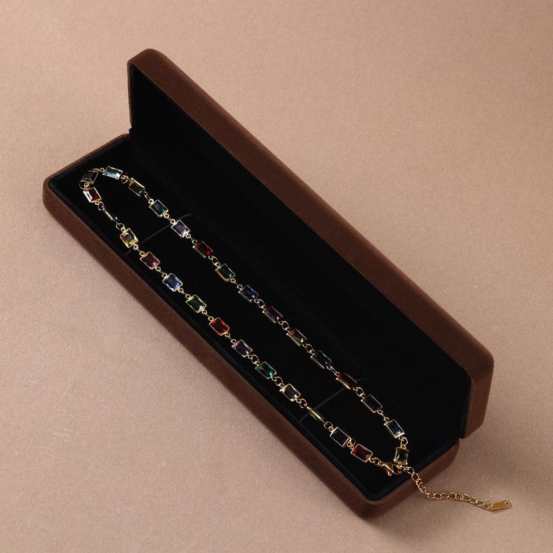 May include: A gold-tone chain necklace featuring a series of rectangular, multi-colored gemstones in shades of red, blue, green, yellow, and purple, presented inside a rectangular brown velvet-lined jewelry box with a hinged lid.