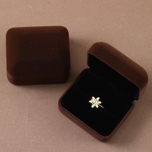 May include: A gold-toned ring featuring a snowflake-shaped centerpiece with a small clear stone at its center, displayed inside an open dark brown velvet jewelry box, with a second closed dark brown velvet jewelry box positioned nearby.