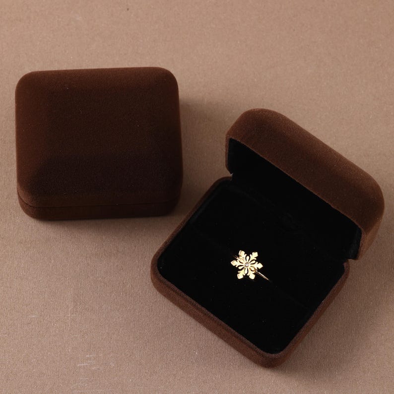 May include: A gold-toned ring featuring a snowflake-shaped centerpiece with a small clear stone at its center, displayed inside an open dark brown velvet jewelry box, with a second closed dark brown velvet jewelry box positioned nearby.