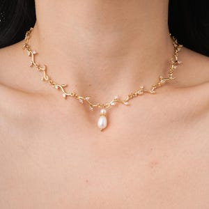 May include: A gold-coloured necklace with a vine-like pattern, adorned with small, clear crystals and a pear-shaped pearl pendant. The necklace is designed to sit around the collarbone, adding a touch of elegance.