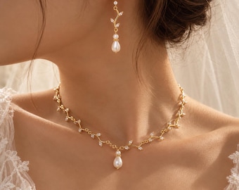 Op de afbeelding: Een goudkleurige ketting en bijpassende oorbellen met een delicaat wijnrankontwerp, versierd met kleine, heldere marquise-geslepen kristallen en een druppelvormige witte parelhangers die in het midden van de ketting hangt.