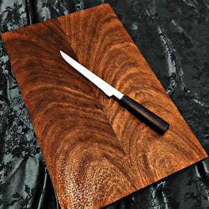 Op de afbeelding: Een rechthoekige houten snijplank met een rijk, donkerbruin nerfpatroon. Een roestvrijstalen mes met een zwart handvat ligt diagonaal over de plank. Het mes heeft een lang, dun lemmet. De achtergrond is een donkere, bedrukte stof.