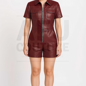 May include: Burgundy-colored leather romper with short sleeves, a collared neckline, and a front zipper. The romper features two chest pockets and a tailored fit. The model is wearing black sandals.