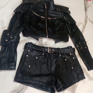May include: A black leather two-piece set consisting of a cropped jacket and matching shorts. The jacket features a high collar, silver-tone zippers, and structured shoulder detailing. The shorts include a belt with a silver buckle and silver studs.