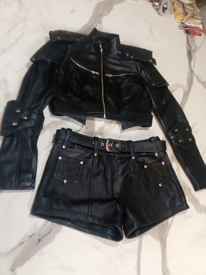 May include: A black leather two-piece set consisting of a cropped jacket and matching shorts. The jacket features a high collar, silver-tone zippers, and structured shoulder detailing. The shorts include a belt with a silver buckle and silver studs.