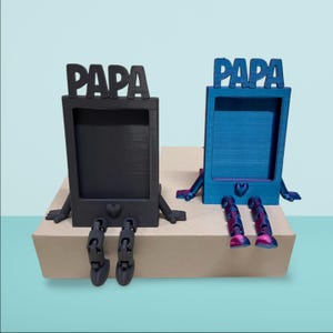 Op de afbeelding: Twee decoratieve fotolijsten, één zwart en één blauw, elk met het woord "PAPA" bovenaan. De lijsten hebben poten en voeten en staan op een beige oppervlak. De blauwe lijst heeft paarse accenten.