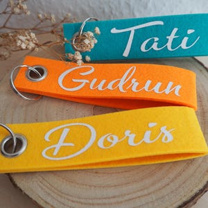 Könnte beinhalten: Drei personalisierte Schlüsselanhänger in Türkis, Orange und Gelb. Jeder Schlüsselanhänger hat einen Namen in weißer Schrift: "Tati", "Gudrun" und "Doris". Die Schlüsselanhänger sind aus Filz und haben einen silbernen Metallring.