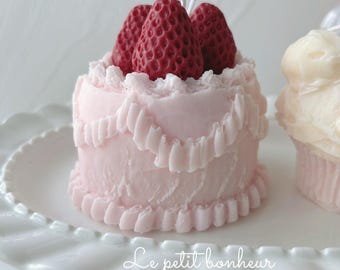 May include: A pink candle shaped like a small cake, topped with three red strawberry candles. The cake has ruffled frosting and sits on a white pedestal. The text "Le petit bonheur" and "Happiness never blows out" are visible.