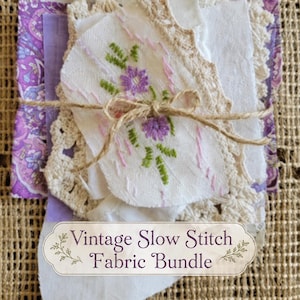 May include: A fabric bundle tied with twine, featuring assorted white lace, cream crochet pieces, and purple floral patterned fabric. One white fabric scrap displays purple and green floral embroidery. A label reads: Vintage Slow Stitch Fabric Bundle.