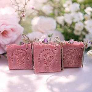 May include: Three square, pink soaps with the text "GREEN WITCH BOTANICALS" and a botanical design. Each soap is tied with twine and topped with a small, purple flower bud. The background features soft-focus pink and white flowers.