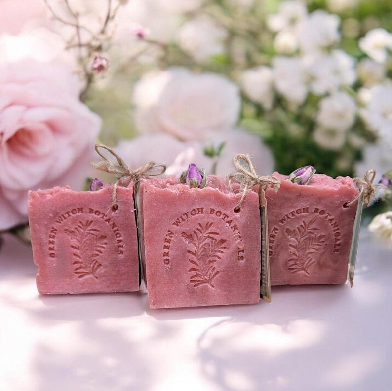 May include: Three square, pink soaps with the text "GREEN WITCH BOTANICALS" and a botanical design. Each soap is tied with twine and topped with a small, purple flower bud. The background features soft-focus pink and white flowers.