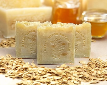 May include: Three square, light-colored soaps with the text "GREEN WITCH BOTANICALS" and a botanical design are in the foreground, surrounded by rolled oats. Jars of honey and more soaps are in the background.