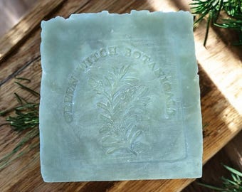 May include: A square bar of light green soap featuring an embossed design of a sprig of rosemary in the center, surrounded by the circular text "GREEN WITCH BOTANICALS" stamped into the surface of the soap.