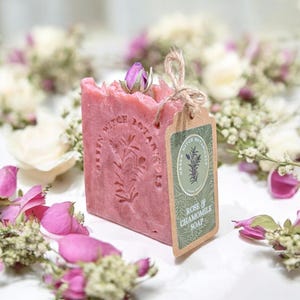 May include: A pink Rose & Chamomile soap bar with a floral design and a small rose bud on top. The soap has a tag that reads "Green Witch Botanicals". The soap is surrounded by flowers.