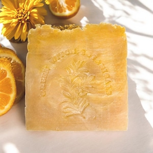 May include: A light orange soap bar with the text "Green Witch Botanicals" and a botanical design. The soap is next to a yellow flower and orange slices.