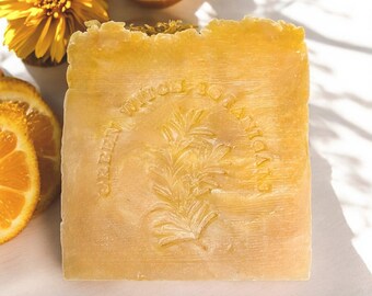May include: A square, light orange soap bar with the text "Green Witch Botanicals" and a botanical design. The soap is next to a yellow flower and orange slices.