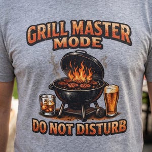 May include: A grey t-shirt with a barbecue-themed graphic. It shows a grill with flames and burgers, a glass of whiskey, and a pint of beer. The text says "GRILL MASTER MODE" and "DO NOT DISTURB" in orange and brown.