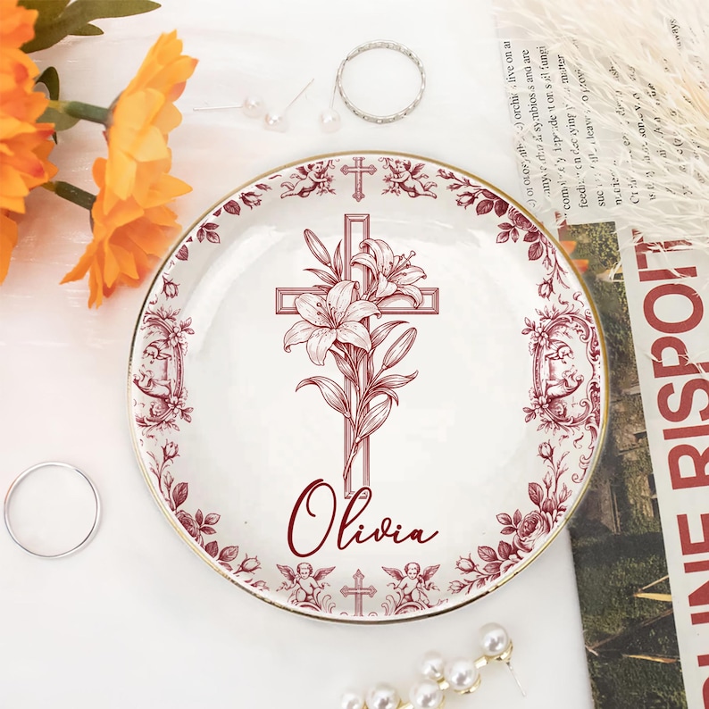 Op de afbeelding: Een witte decoratieve plaat met een bordeaux kruis- en lelie-ontwerp, gepersonaliseerd met de naam "Olivia". De plaat heeft een gouden rand en een bloemenrand. Extra ringen en bloemen zijn op de achtergrond.
