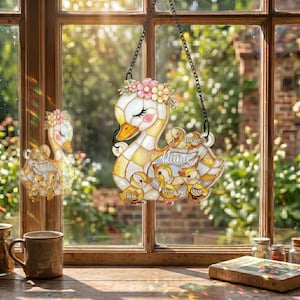 May include: A decorative swan-shaped hanging ornament with a floral crown and the word "Nana". The ornament features a mother swan with several ducklings, each with a name. The design is in shades of white, yellow, and gold, with a black chain for hanging.
