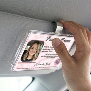 Puede incluir: Un clip de visera de coche de pl&aacute;stico transparente que sostiene una tarjeta de identificaci&oacute;n de princesa pasajera rosa y blanca. La tarjeta de identificaci&oacute;n presenta una foto de una mujer sonriente y el texto "Pas Princess".