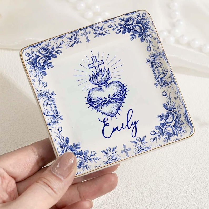 Op de afbeelding: Vierkante keramische sieradenschaal met een blauwe bloemenrand en gouden rand. Het midden toont een blauw hart met een doornenkroon, een kruis en de naam "Emily". Decoratief en gepersonaliseerd.