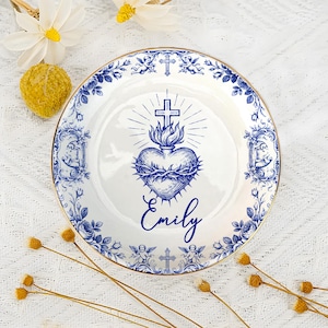 Op de afbeelding: Witte keramische plaat met een blauwe bloemenrand en een gouden rand. De plaat heeft een blauw hartontwerp met een kruis en de naam "Emily" in script. Decoratief en sierlijk.