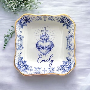 Op de afbeelding: Vierkante keramische schaal met een gouden rand en een blauw-wit bloemmotief. Het midden heeft een blauw hart met een kruis en de naam "Emily". Decoratieve cherubs en kruisen zijn ook aanwezig.